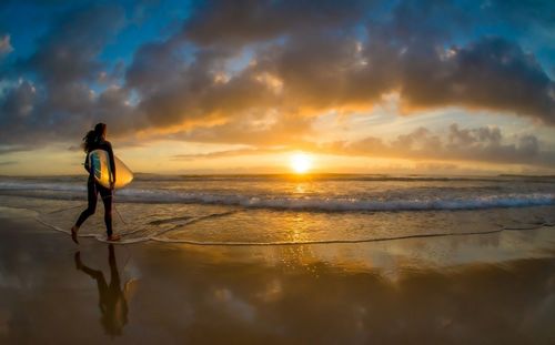 Full length of woman with surfboard walking on beach during sunset