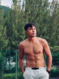 Young man looking away while standing against trees