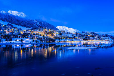 Illuminated buildings by lake against blue sky during winter