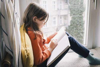 Side view of girl reading book while sitting on window sill at home