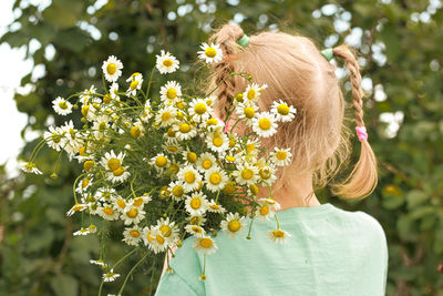 A girl with blond hair in pigtails stands with her back with a bouquet of daisies over her shoulder