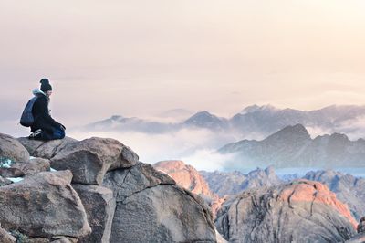 Man sitting on rock looking at mountains against sky