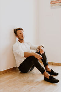 Full length of man looking away while sitting on hardwood floor against wall
