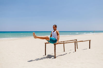 Man exercising on parallel bars at beach against blue sky