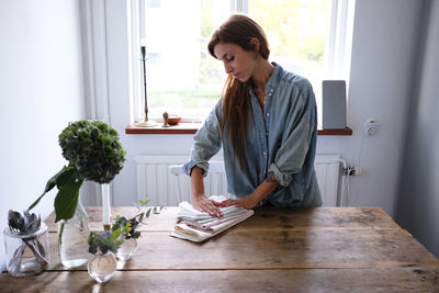 Mid adult woman folding napkins while standing at wooden table in kitchen