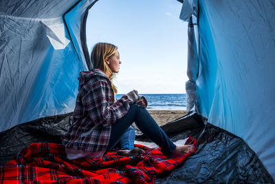 Side view of young woman sitting in tent at beach