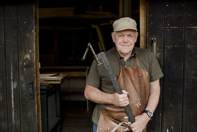 Portrait of confident senior male owner holding equipment while standing at store entrance