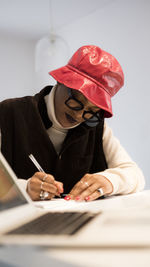 Woman wearing hat writing while sitting at desk in office