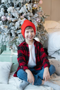 A boy in a red cap and a plaid shirt sits near a christmas tree with gifts on the floor and smiles