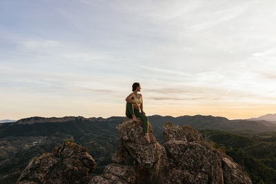 Young confident barefoot female in activewear relaxing on rocky cliff while having yoga practice at sunrise