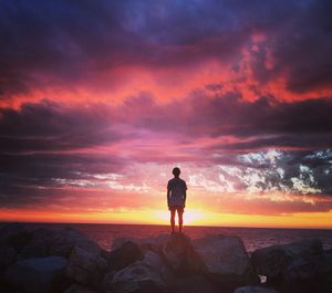 Rear view of young man standing on rocks at seashore against dramatic sky