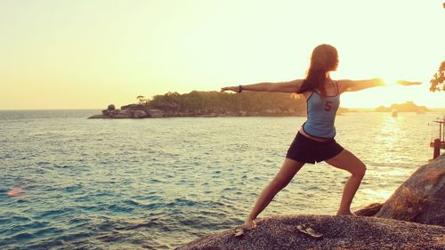 Woman performing warrior 2 on beach against clear sky at sunset