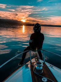 Rear view of man sitting on railing of boat in sea against sky during sunset