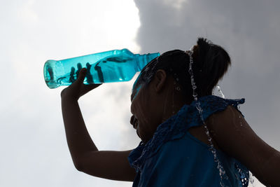 Silhouette of a girl splashing water on head from a blue plastic bottle under sky to cool down