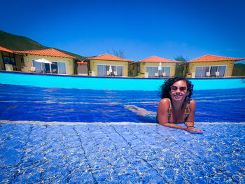 Full length of young woman sitting in swimming pool against blue sky