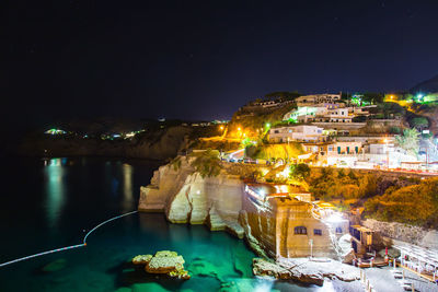 Illuminated city by sea against sky at night