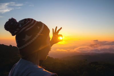 Woman gesturing ok sign against sky during sunset