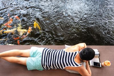 Rear view of man reading book while lying on pier over lake