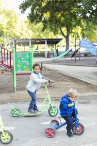 Boy with push scooter looking at friend riding tricycle on playground