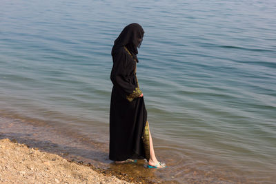 A muslim woman in national clothes wets her feet in sea.