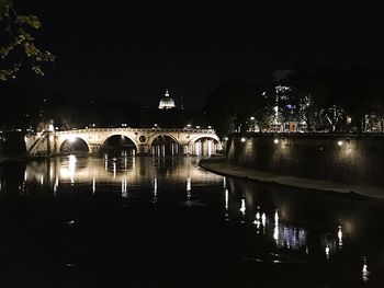 Bridge over river by illuminated buildings against sky at night