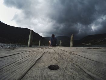 Rear view of woman sitting on boardwalk against cloudy sky