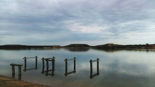 Wooden posts in lake by mountains against cloudy sky
