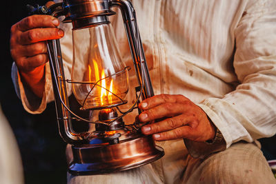 Man in white untidy cotton working suit holding large lit kerosene lamp in his hands