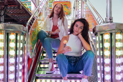 Young female friends in casual clothes with candies in hands looking at camera while sitting on steps of attraction in funfair in summer evening