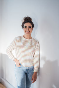 Smiling woman with hand in pocket leaning on white wall at home