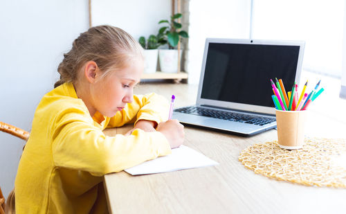 Schoolgirl in a yellow sweatshirt is doing homework at home by the window.
