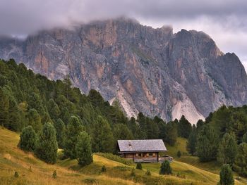 A wooden house in a meadow next to a forest under a rocky mountain in the dolomites, italy