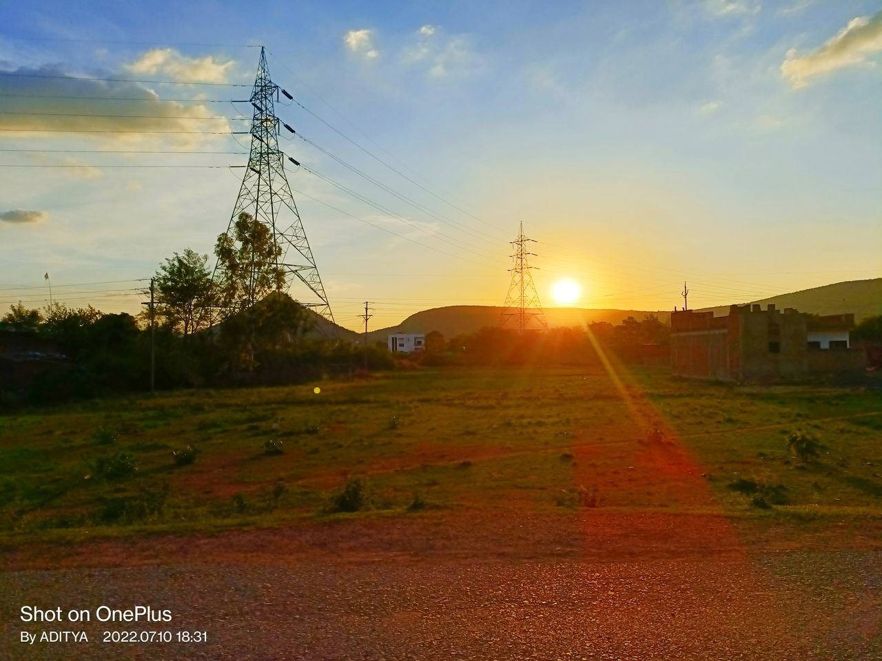 sky, sunset, horizon, sunlight, nature, sun, cloud, electricity, field, landscape, technology, evening, environment, electricity pylon, dusk, lens flare, plain, plant, architecture, cable, no people, tree, sunbeam, rural area, built structure, land, beauty in nature, power generation, hill, grass, power line, scenics - nature, power supply, transportation, outdoors, orange color, afterglow, tranquility, road, rural scene, agriculture