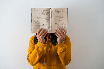 Woman holding book in front of face against white wall