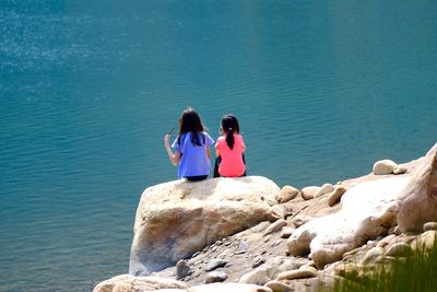 Rear view of girls sitting on rock formation by lake