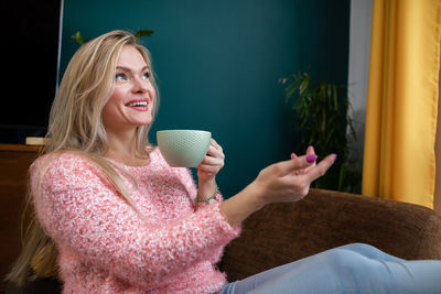 A young woman laughs while drinking coffee from a cup at home.