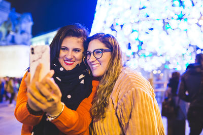 Smiling young women taking selfie while standing against illuminated lighting outdoors