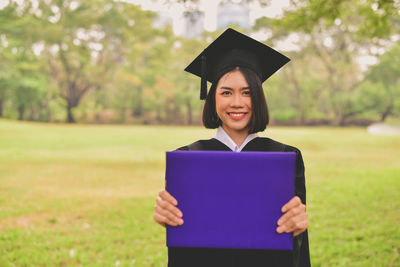 Portrait of young woman in graduation gown holding file while standing at park