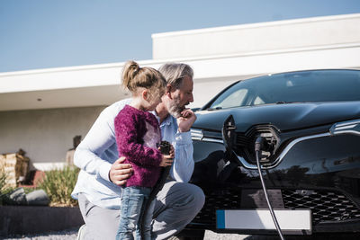 Girl holding electric plug standing by father looking at car on sunny day