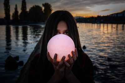 Portrait of girl holding crystal ball against lake during sunset