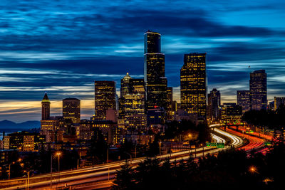 Light trail on street by illuminated buildings in city against sky