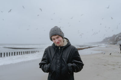 A guy with a grimace on his face in a winter hat and a black jacket stands on the seashore