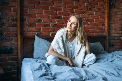 Young adult woman with blonde long hair in casual clothes sitting on bed at home
