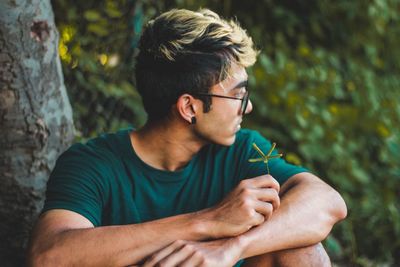Young man looking away while sitting by tree trunk