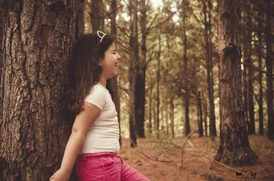 Side view of young woman standing by tree trunk in forest