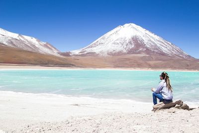 Man sitting on shore against mountains against clear blue sky