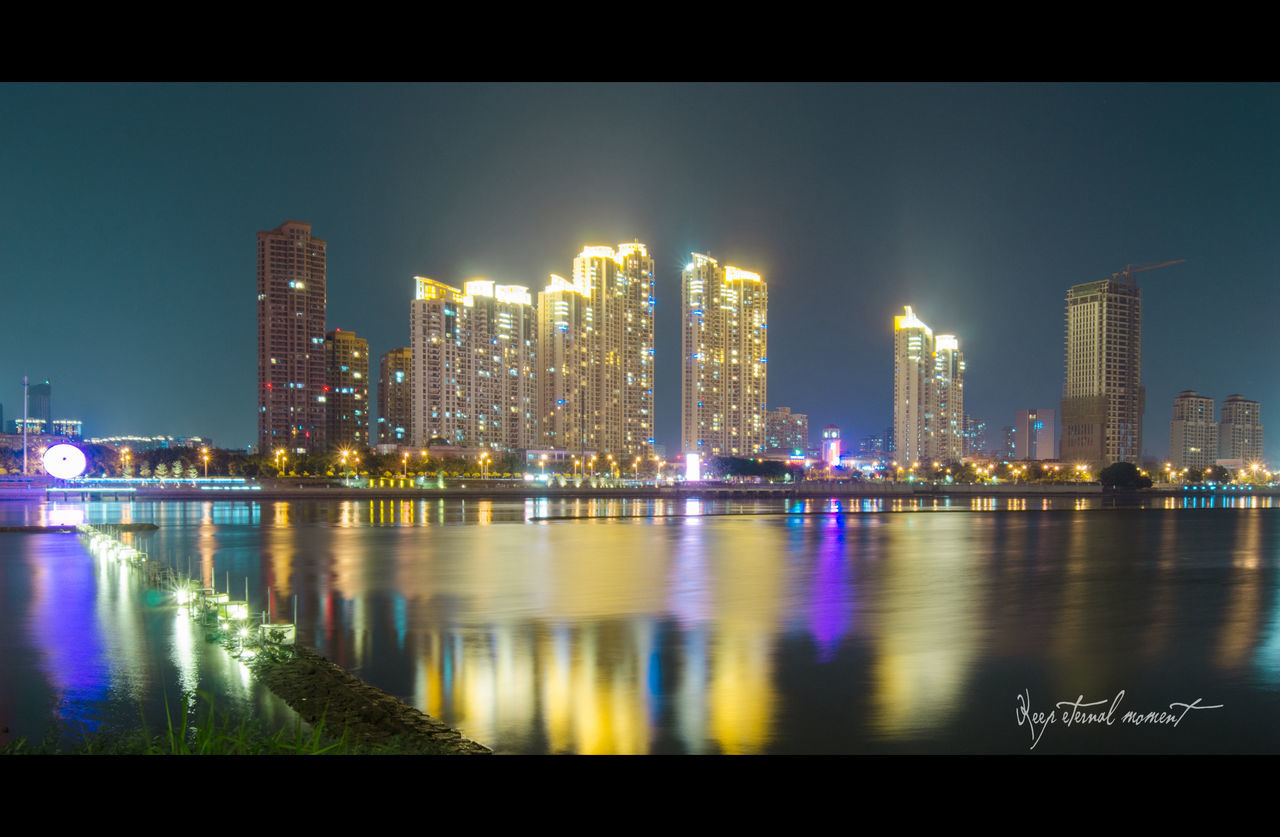 illuminated, water, building exterior, night, city, reflection, architecture, built structure, transfer print, cityscape, waterfront, skyscraper, river, auto post production filter, urban skyline, sky, modern, clear sky, sea, blue