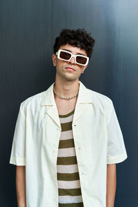 Confident handsome male with curly hair dressed in white shirt and trendy sunglasses looking at camera on gray background