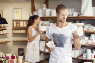 Male worker looking at craft products while female colleague working in background in crockery workshop