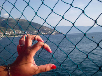 Cropped image of hand by sea against sky seen through chainlink fence